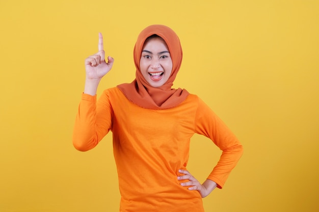 Sonriente mujer asiática feliz con su dedo apuntando hacia arriba aislado sobre fondo de banner amarillo claro con hijab