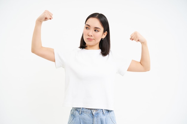 Sonriente mujer asiática flexionando bíceps luciendo atrevida y fuerte mostrando su fuerza músculos studi blanco