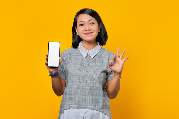 Sonriente mujer asiática que muestra la pantalla en blanco del teléfono inteligente y gesticula bien firmar sobre fondo amarillo