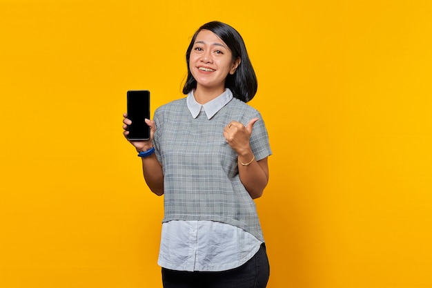 Sonriente mujer asiática que muestra la pantalla del teléfono inteligente en blanco y los pulgares para arriba sobre fondo amarillo