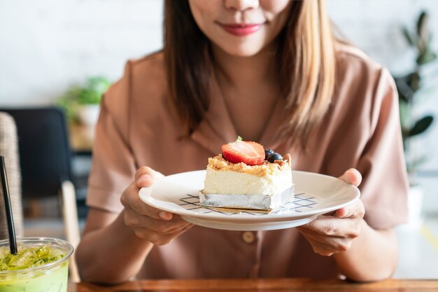 Sonriente mujer asiática sosteniendo un plato de su tarta de queso de fresa favorita en la mesa de madera en la cafetería.