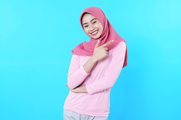 Sonriente mujer asiática con su dedo apuntando aislado sobre fondo azul claro vistiendo hijab con camiseta rosa