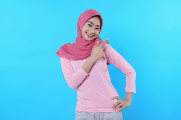 Sonriente mujer asiática con su dedo apuntando aislado sobre fondo azul claro vistiendo hijab con camiseta rosa