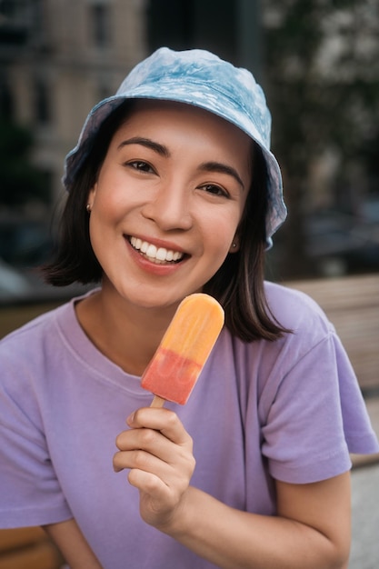 Sonriente mujer asiática vistiendo panama comiendo helado mirando a la cámara. postre sabroso