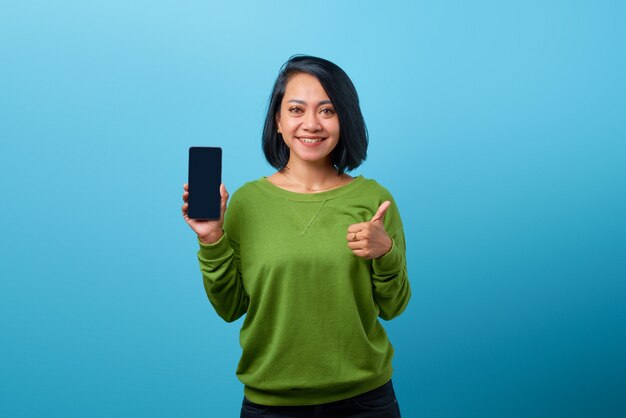 Sonriente mujer asiática vistiendo ropa casual sosteniendo smartphone y mostrando los pulgares para arriba