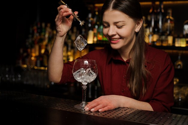 Sonriente mujer barman poniendo cubitos de hielo en un vaso haciendo un cóctel de jeringas Aperol