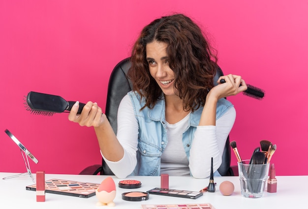 Foto sonriente mujer bastante caucásica sentada a la mesa con herramientas de maquillaje sosteniendo peines y apuntando al lado