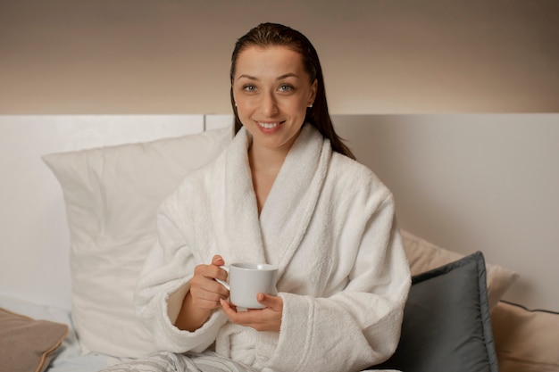 Sonriente mujer en bata de baño sentado en la cama bebiendo té de hierbas caliente