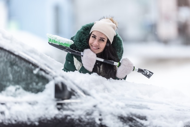 Sonriente mujer bien vestida sostiene un raspador de hielo y una escoba de nieve mientras se inclina contra el coche cubierto de nieve.