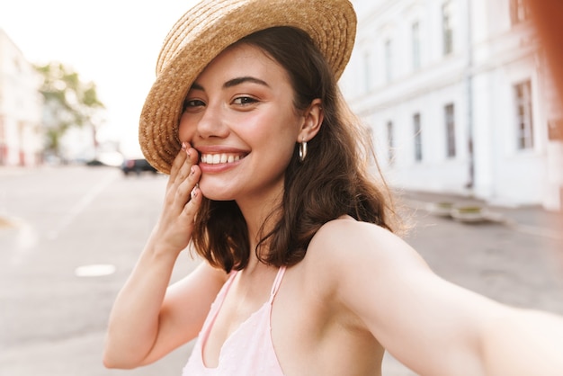 Sonriente mujer bonita joven alegre positiva con hermoso sombrero caminando por la calle tomar selfie por cámara.