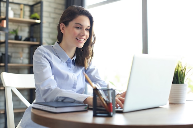 Sonriente mujer bonita sentada a la mesa, mirando la pantalla del portátil. Feliz empresario leyendo un mensaje de correo electrónico con buenas noticias, charlando con clientes en línea.