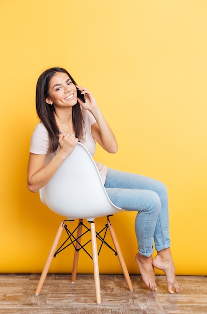 Sonriente mujer bonita sentada en la silla y hablando por teléfono sobre pared amarilla