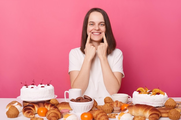 Sonriente mujer de cabello castaño con camiseta blanca sentada en la mesa con sabrosos dulces apuntando a sus dientes cuidado dental aislado sobre fondo rosa