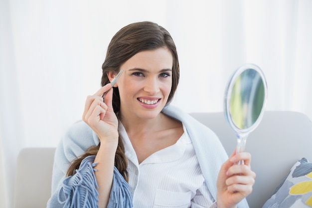 Sonriente mujer de cabello castaño casual en pijama blanco desplumando las cejas