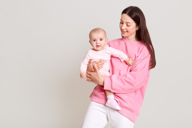 Sonriente mujer de cabello oscuro en ropa casual sosteniendo a la pequeña niña en las manos, mira a su bebé, niño emocionado con traje, posando aislado sobre una pared blanca.