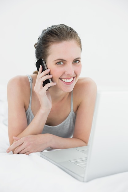 Sonriente mujer casual con teléfono celular y computadora portátil en la cama