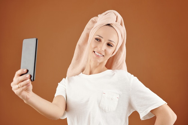 Sonriente mujer caucásica bastante joven con una toalla en la cabeza tomando selfie en smartphone contra fondo marrón