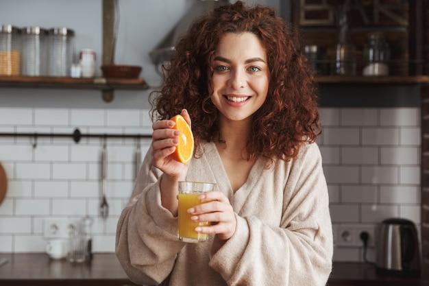 Sonriente mujer caucásica bebiendo jugo de naranja fresco mientras desayuna en el interior de la cocina de casa