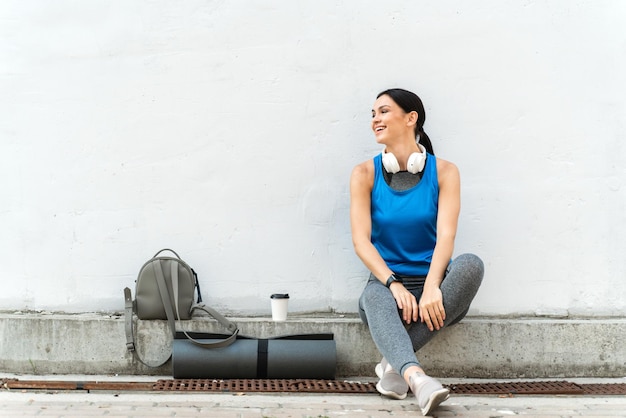 Foto sonriente mujer caucásica en forma animada sentada con las piernas cruzadas por un muro de hormigón