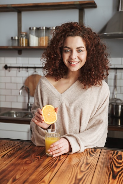 Sonriente mujer caucásica haciendo y bebiendo jugo de naranja fresco mientras desayuna en el interior de la cocina de casa