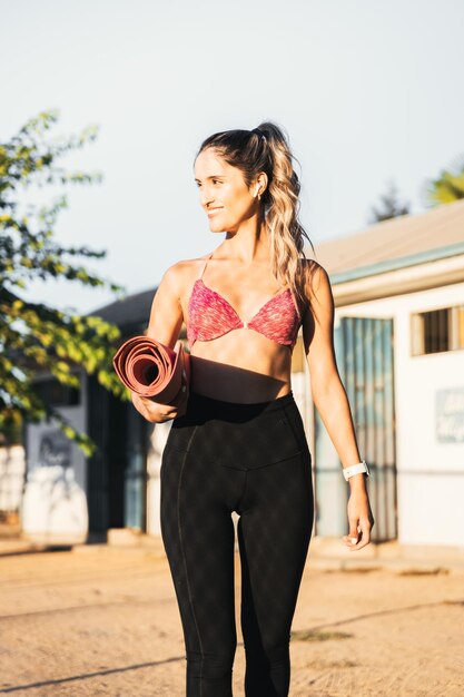 Sonriente mujer caucásica joven deportiva activa caminando al frente y sosteniendo su estera de yoga rosa