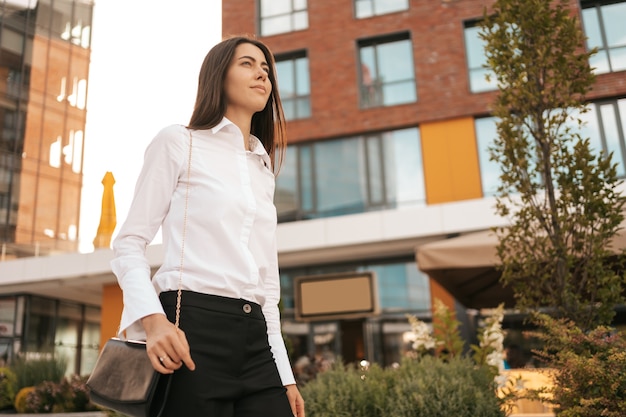 Sonriente mujer caucásica en ropa de negocios caminando cerca del centro de negocios urbano