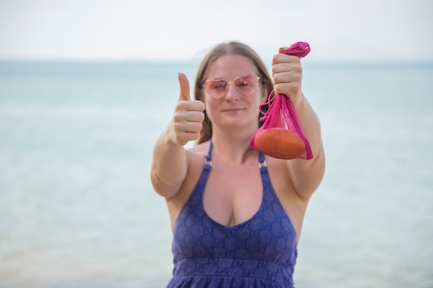 Sonriente mujer caucásica sosteniendo una bolsa de malla con frutas y mostrando los pulgares para arriba en el mar o el océano