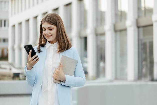 Sonriente mujer caucásica con traje formal usando teléfono móvil escribiendo mensajes de texto caminando sosteniendo