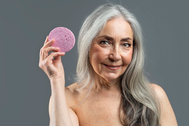 Sonriente mujer desnuda europea jubilada con cabello gris muestra esponja de masaje para la cara