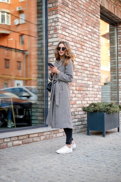 Sonriente mujer elegante en gabardina caminando y bebiendo café de la taza de papel. mujer sonriente con el pelo rizado mira a cámara sonrisa caminando cámara lenta cara puesta de sol hermosa dama al aire libre primer plano lindo