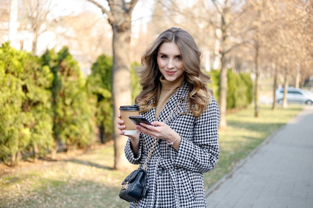 Sonriente mujer elegante en gabardina caminando sosteniendo móvil y café