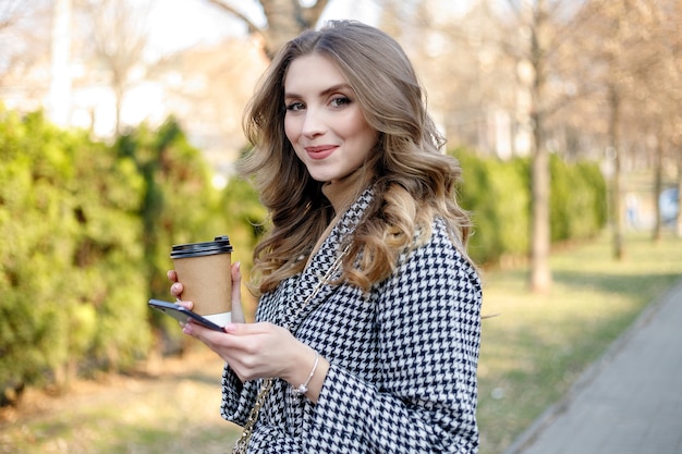 Sonriente mujer elegante en gabardina caminando sosteniendo móvil y café