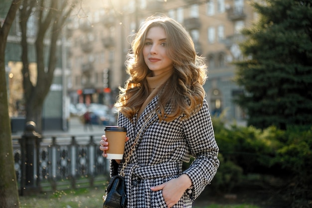 Sonriente mujer elegante en gabardina caminando y tomando café de la taza de papel. mujer sonriente con el pelo rizado mira a cámara sonrisa caminando cámara lenta cara puesta de sol hermosa dama al aire libre primer plano lindo