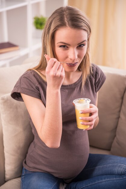 Sonriente mujer embarazada está comiendo mientras está sentado en el sofá.