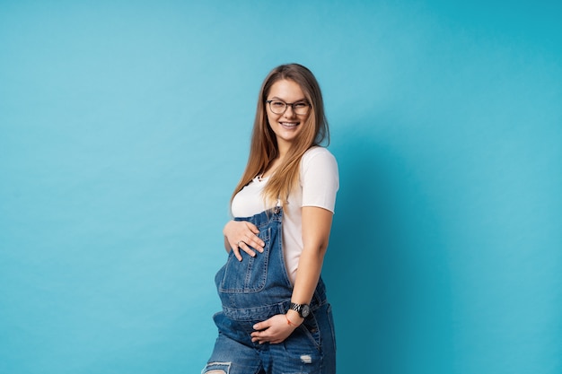 Sonriente mujer embarazada con gafas acariciando su vientre sobre fondo azul.