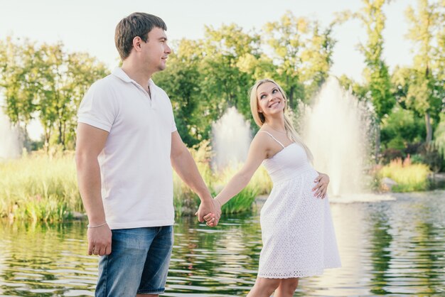 Sonriente mujer embarazada mirando a su marido cerca del lago en la noche cogidos de la mano. Tiempo libre al aire libre.