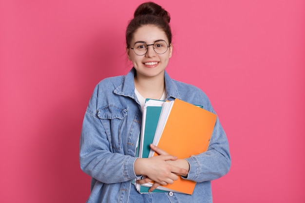 Sonriente mujer estudiante con cabello oscuro y nudo, sosteniendo carpetas de papel