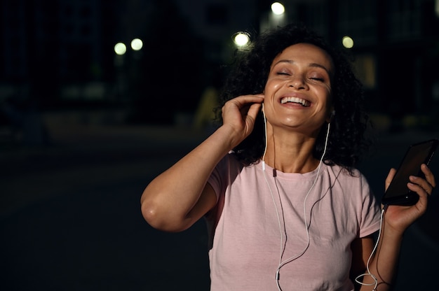 Sonriente mujer de etnia afroamericana con auriculares, sosteniendo un teléfono móvil y disfrutando de escuchar la música con los ojos cerrados