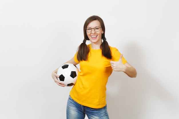 Sonriente mujer europea, dos divertidas colas de caballo, aficionado al fútbol o jugador con gafas, uniforme amarillo muestran el pulgar hacia arriba, sostienen el balón de fútbol aislado sobre fondo blanco. Deporte, fútbol, concepto de estilo de vida saludable.