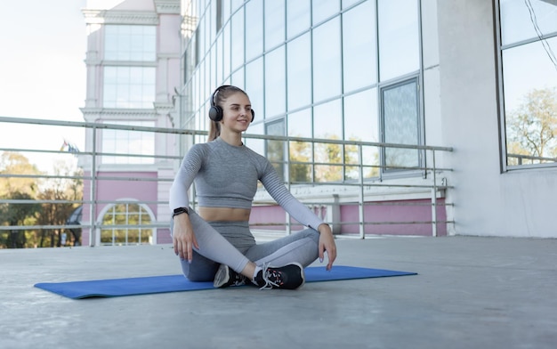 Sonriente mujer europea en forma sentada en una alfombra y escuchando música en el techo del edificio