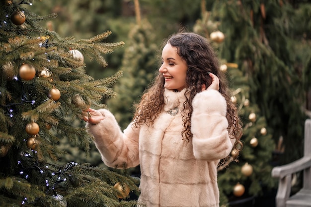 Sonriente mujer feliz de cabello rizado en un abrigo de pieles pasea por la feria del árbol de Navidad Esperando Navidad y Año Nuevo