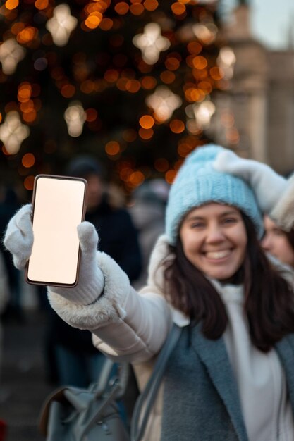Sonriente mujer feliz en la feria de la ciudad de navidad sosteniendo el teléfono con pantalla blanca