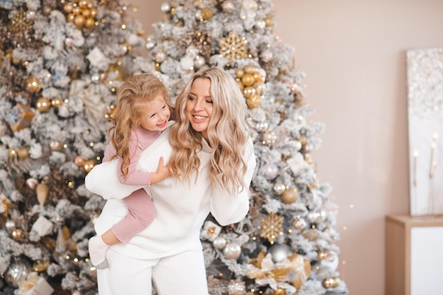 Sonriente mujer feliz sosteniendo a una niña jugando sobre un árbol de Navidad decorado en la habitación Temporada de vacaciones de invierno Maternidad