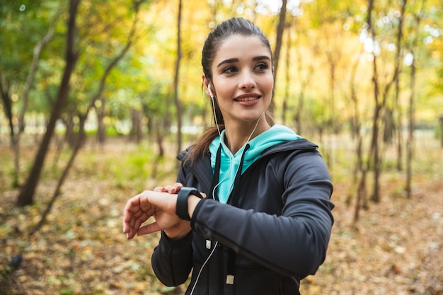 Sonriente mujer fitness escuchando música con auriculares, comprobando smartwatch, mientras está de pie en el parque