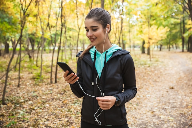 Sonriente mujer fitness escuchando música con auriculares, sosteniendo el teléfono móvil mientras está de pie en el parque