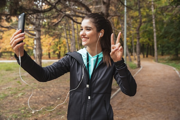 Sonriente mujer fitness escuchando música con auriculares, tomando un selfie mientras está de pie en el parque