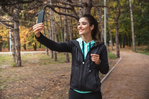 Sonriente mujer fitness escuchando música con auriculares, tomando un selfie mientras está de pie en el parque