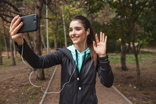 Foto sonriente mujer fitness escuchando música con auriculares, tomando un selfie mientras está de pie en el parque