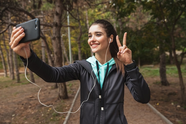 Sonriente mujer fitness escuchando música con auriculares, tomando un selfie mientras está de pie en el parque