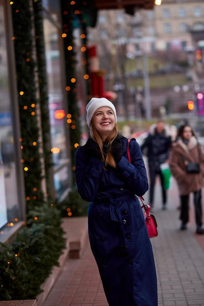 Sonriente mujer hermosa en la calle de la noche cerca de escaparate decorado Elegante ropa casual de invierno Luces de la ciudad Ciudad festiva